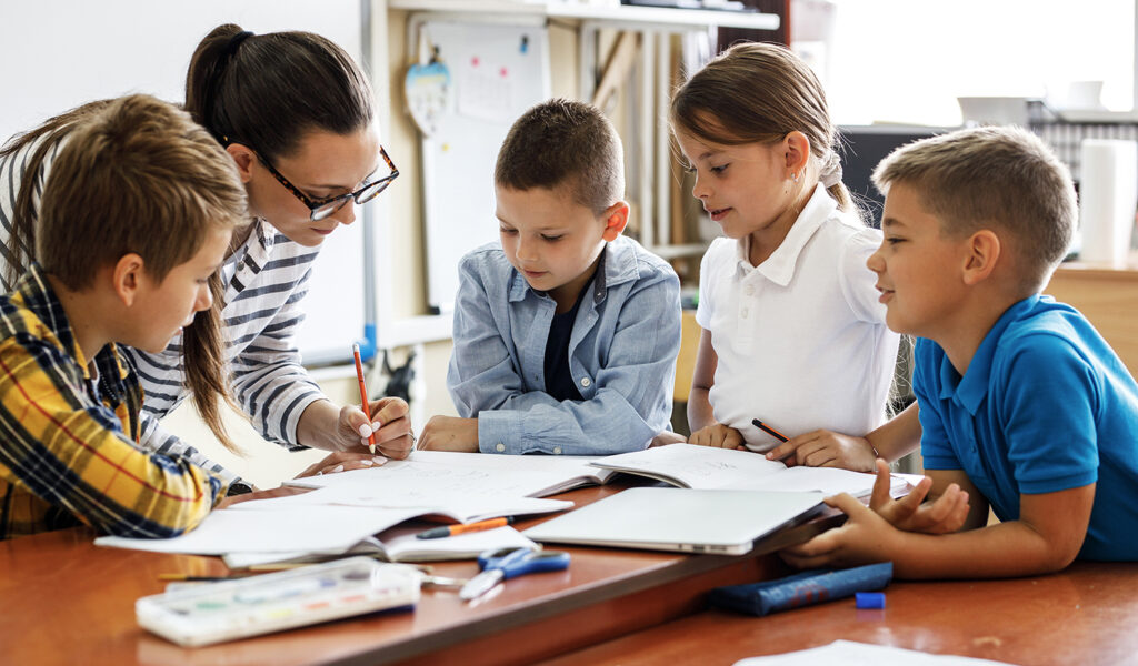 Eine Lehrerin oder Betreuerin gibt Grundschulkindern in Ludwigsburg Hausaufgabenhilfe an einem großen Schreibtisch.